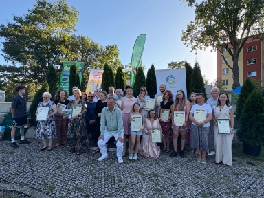 Grupa około 25 osób na zewnątrz, w dzień, trzymających dyplomy, stojących i kucających na tle zielonych drzewek i kilku banerów reklamowych.