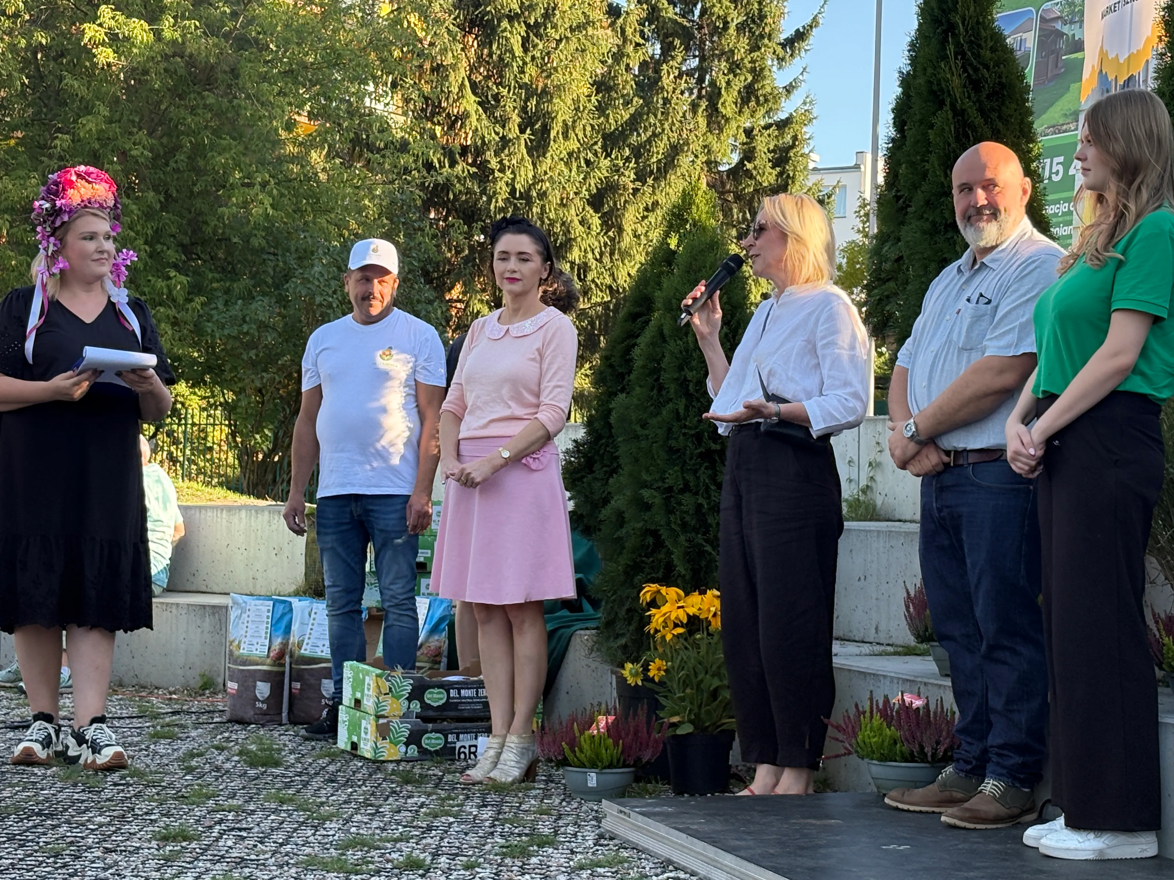 Sześć osób stoi na zewnątrz na małej scenie i obok niej, jedna kobieta mówi do mikrofonu, na pierwszym planie kwiaty i torby z ziemią, za nimi drzewa, dzień.