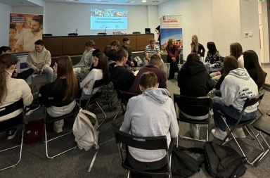 Grupa około dwudziestu młodych osób siedzi na krzesłach ustawionych w dwóch kołach w sali konferencyjnej z jasnymi ścianami i dywanem na podłodze. Na ścianie w tle znajduje się ekran z wyświetlanym tekstem oraz dwa stojące banery. Uczestnicy rozmawiają ze sobą, większość ma casualowy strój, niektórzy z nich mają plecaki obok krzeseł.