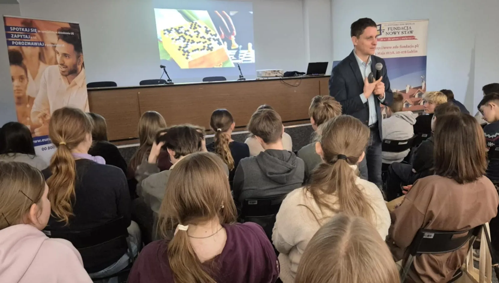 Grupa młodych osób siedzi w sali wykładowej zwrócona w stronę mężczyzny prowadzącego prezentację z mikrofonem. Na ścianie za nim wyświetlony jest obraz gry planszowej. Po bokach stoją dwa stojące banery informacyjne.