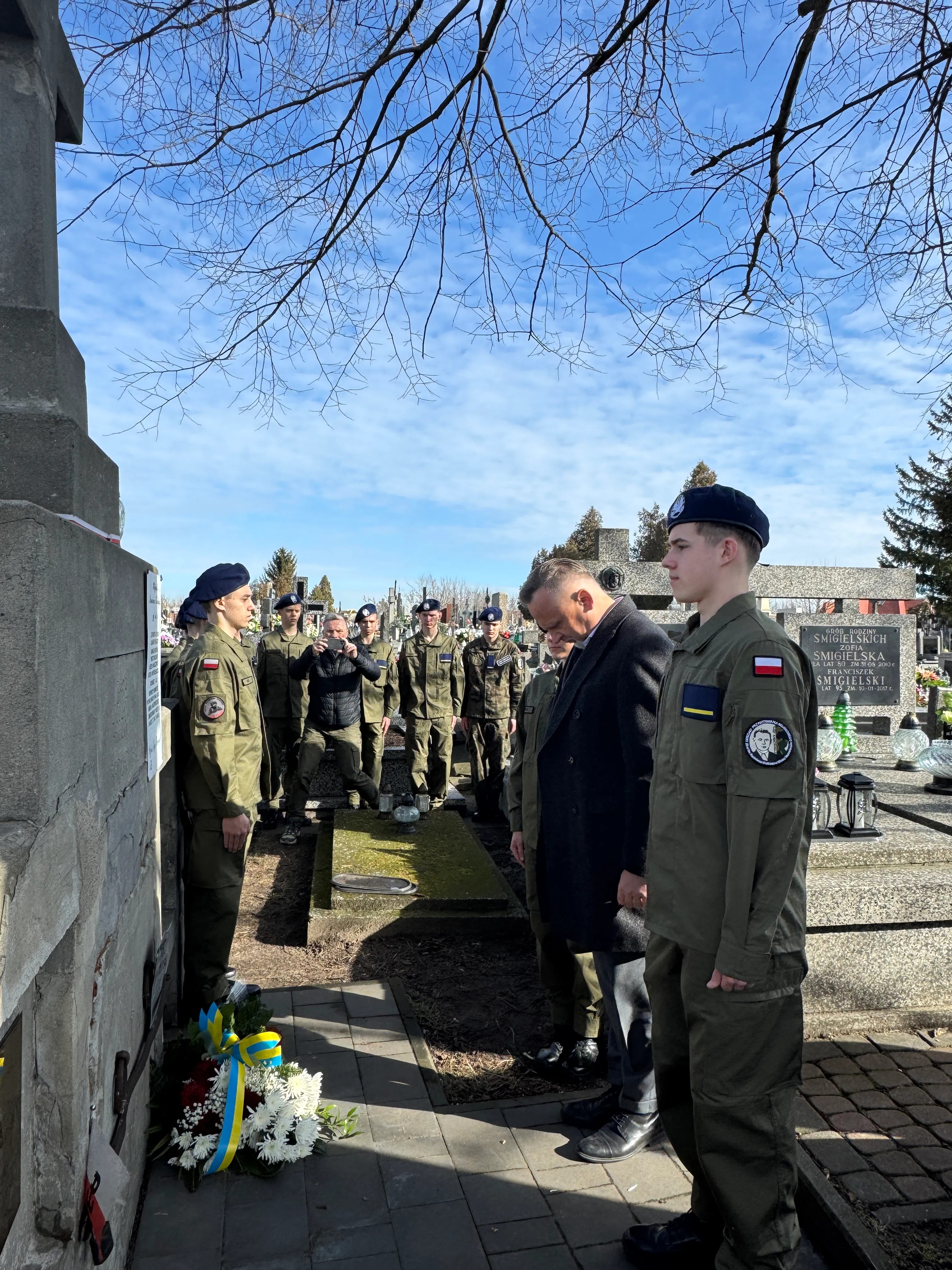 Grupa żołnierzy i mężczyzna w ciemnym płaszczu stoją na cmentarzu przy nagrobku. Na chodniku leży wieniec z niebiesko‑żółtą wstążką, w tle nagie gałęzie drzew i błękitne niebo.