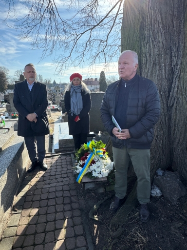 Trzech dorosłych stoi przy dużym drzewie na cmentarzu w słoneczny, zimowy dzień. Mężczyzna po prawej trzyma notes i przemawia. Po lewej mężczyzna w ciemnym płaszczu trzyma kapelusz. Między nimi stoi kobieta w czerwonym berecie i szaliku. U podstawy drzewa leży wieniec z białych i czerwonych kwiatów z niebiesko‑żółtą wstążką. W tle rzędy nagrobków i jasne niebo.