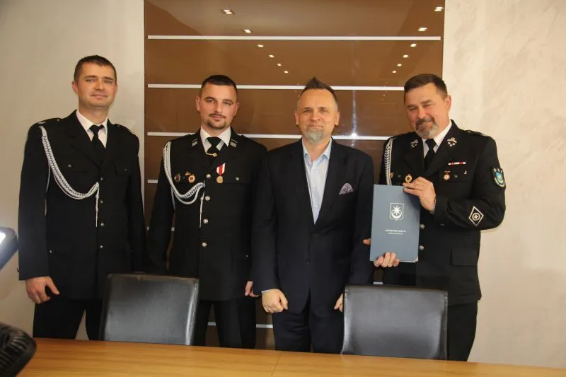 Czterech mężczyzn stoi obok stołu, trzech w mundurach z odznaczeniami, a jeden w ciemnym garniturze, z jednym z mundurowych trzymającym teczkę.