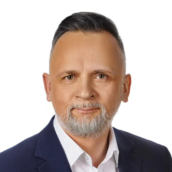 Mężczyzna w średnim wieku z siwą brodą i krótkimi czarnymi włosami ubrany w białą koszulę i granatową marynarkę.