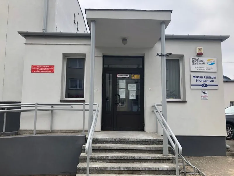 Kliknięcie powiększy obraz. Wejście do budynku z podjazdem i schodami oraz tablicami informacyjnymi przy drzwiach.