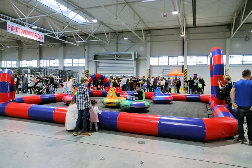 Dzieci i dorośli przy torze do pojazdów bumper car w hali ze stoiskiem oznaczonym jako "Punkt odbioru paczek".