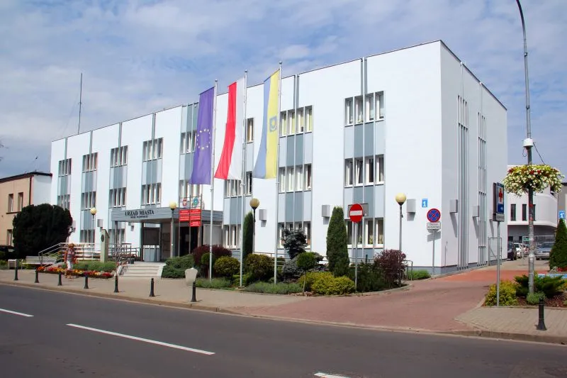 Kliknięcie powiększy obraz. Trzypiętrowy biały budynek z prostokątnymi oknami, czterema flagami na masztach i wejściem oznaczonym napisem „URZĄD MIASTA”.
