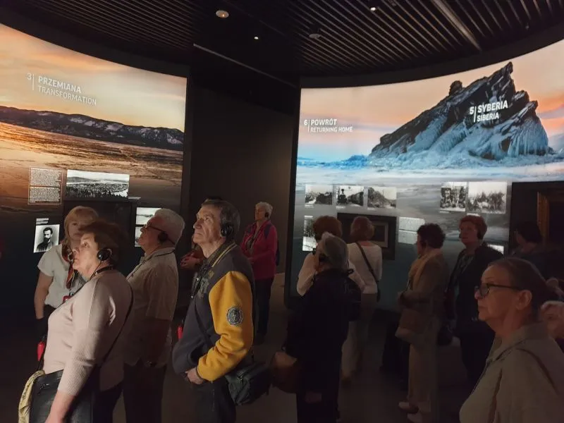 Grupa osób słuchająca przewodnika w muzeum stojąca przed ekspozycjami o transformacji, Syberii i powrocie do domu.