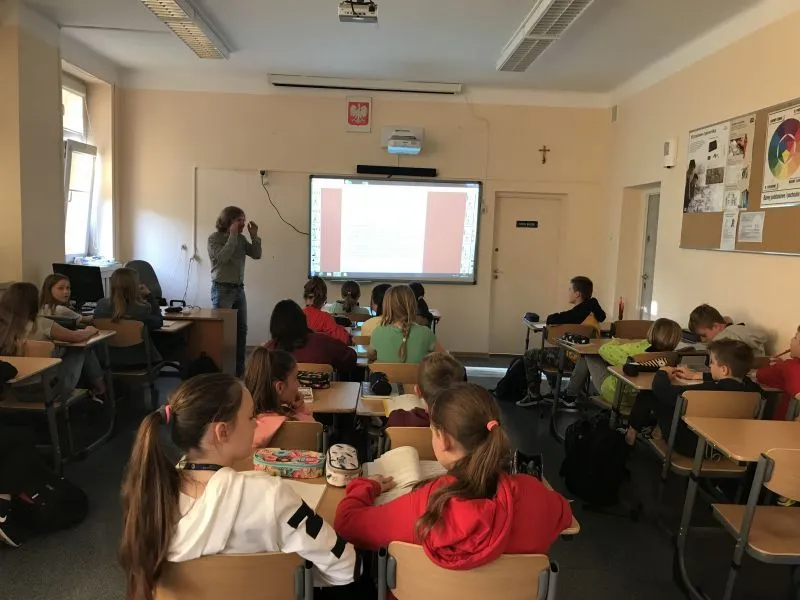 Uczniowie siedzący przy ławkach zwróceni w stronę nauczyciela objaśniającego materiał wyświetlany na tablicy multimedialnej.