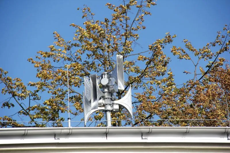 Kliknięcie powiększy obraz. Metalowa wieża syren alarmowych umieszczona na krawędzi dachu budynku na tle drzewa i błękitnego nieba.