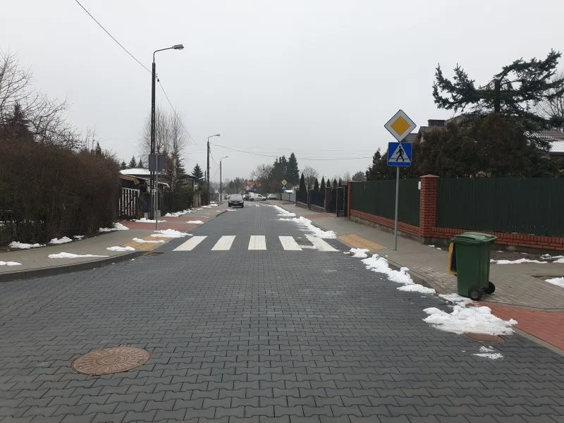 Ulica z przejściem dla pieszych, latarniami, znakami drogowymi oraz rozproszonymi resztkami śniegu na chodnikach i poboczach.