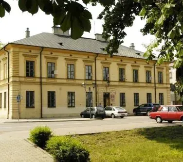 Dwukondygnacyjny żółty budynek z poddaszem, otoczony zielenią i ulicą z zaparkowanymi samochodami.