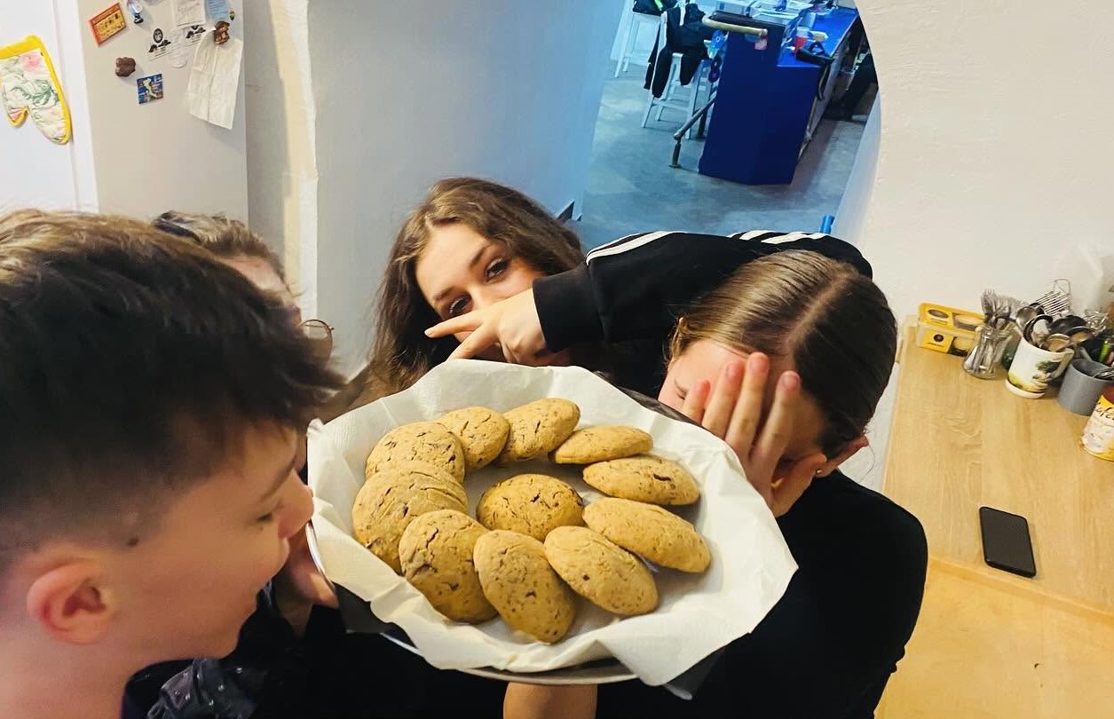 Trzech nastolatków stoi przy kuchennym blacie wewnątrz mieszkania. Jeden trzyma talerz z papilotą pełen dużych okrągłych ciastek ustawionych w kręgu. Jeden nastolatek zasłania twarz dłonią, drugi robi znak V. Na blacie widać przybory kuchenne i telefon.