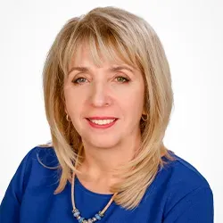 Kobieta w średnim wieku z krótkimi blond włosami, uśmiechnięta, ubrana w niebieską bluzkę i naszyjnik.