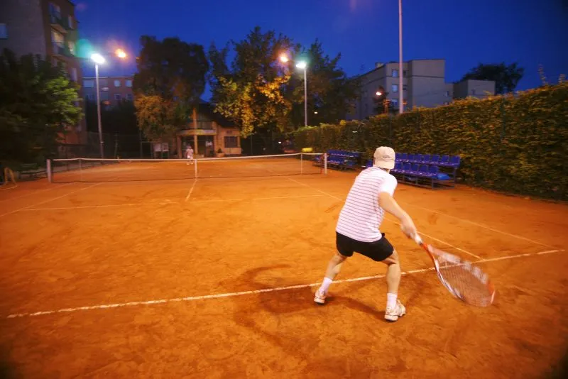 Mężczyzna w czapce i krótkich spodenkach gra w tenisa na oświetlonym korcie ziemnym nocą.