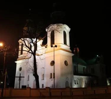 Biały budynek kościoła z dwoma wieżami oświetlony nocą z drzewem i ogrodzeniem na pierwszym planie.