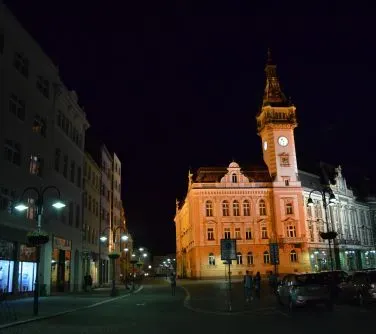 Oświetlony budynek z wysoką wieżą z zegarem nocą na tle ciemnego nieba, otoczony ulicą i zaparkowanymi samochodami.