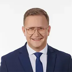 Mężczyzna w okularach, granatowym garniturze, białej koszuli i granatowym krawacie na białym tle.