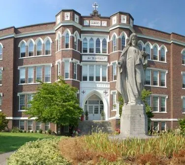 Duży budynek z czerwonej cegły o gotyckich oknach i wejściu oraz kamienna statua postaci z uniesioną dłonią na pierwszym planie.