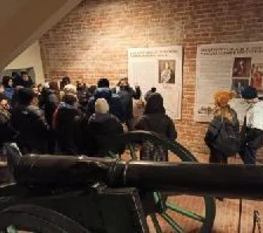 Grupa osób oglądająca wystawę na ceglonej ścianie muzealnej z widocznym przed nimi działem artyleryjskim.