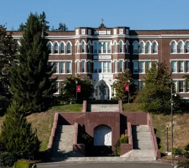 Trzypiętrowy budynek z czerwonej cegły z wieloma białymi oknami i centralnym wejściem z łukiem oraz schodami prowadzącymi do niego przez zieloną przestrzeń.