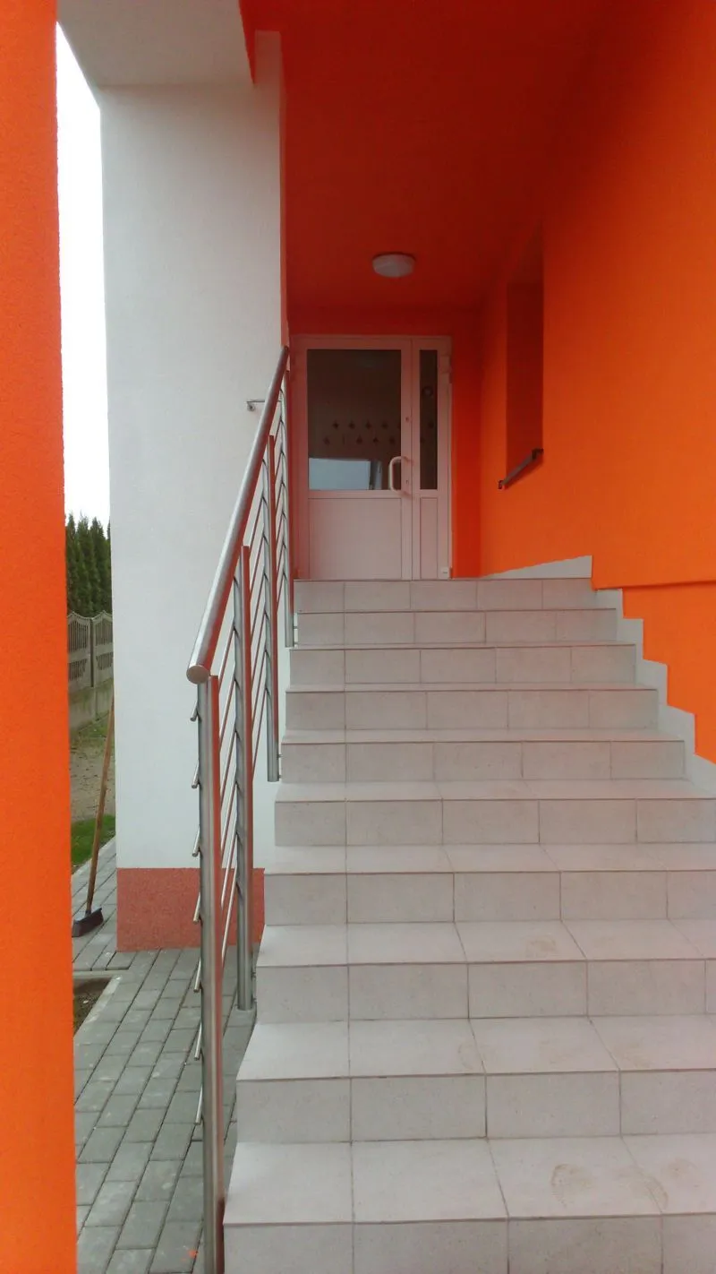 Schody z jasnych płytek z metalową poręczą prowadzące do białych drzwi na tle pomarańczowej ściany.