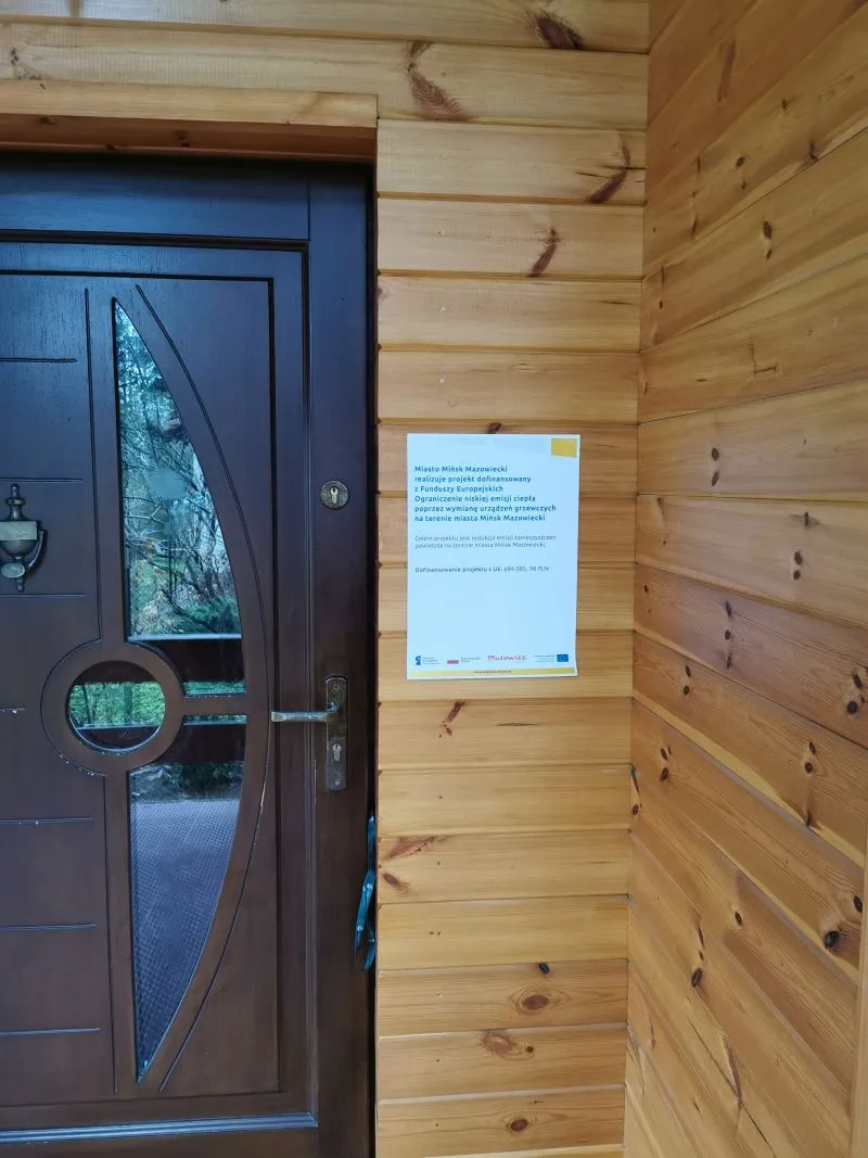 Brązowe drewniane drzwi z ozdobnym szkłem obok drewniana ściana z zawieszoną białą tabliczką informacyjną.