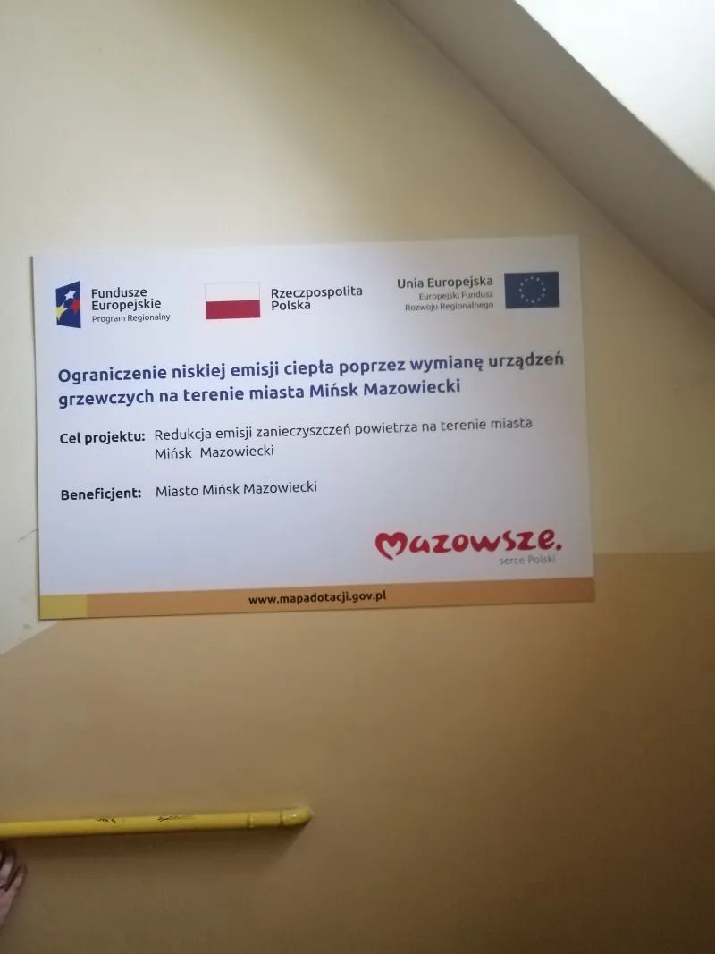 Tablica informacyjna o projekcie ograniczenia niskiej emisji ciepła poprzez wymianę urządzeń grzewczych na terenie miasta Mińsk Mazowiecki z logotypami Funduszy Europejskich, Rzeczpospolitej Polskiej i Unii Europejskiej.