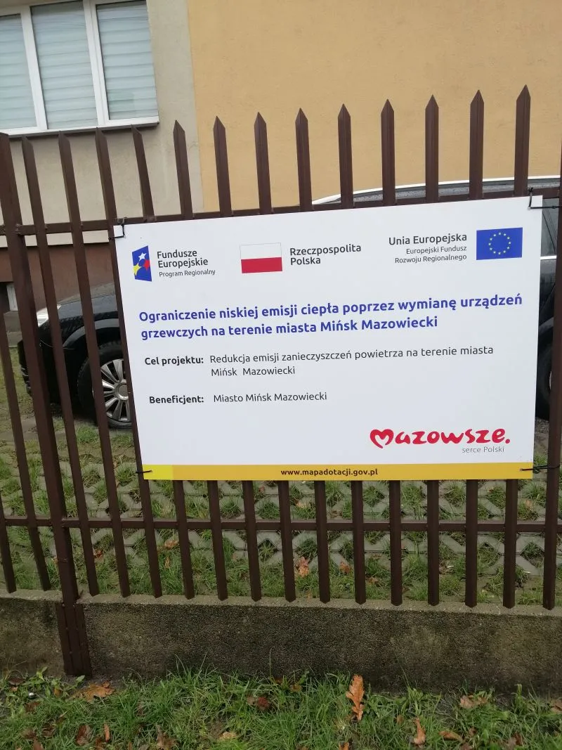Tablica informacyjna o projekcie ograniczającym niską emisję ciepła poprzez wymianę urządzeń grzewczych w Mińsku Mazowieckim z logotypami Funduszy Europejskich, Rzeczpospolitej Polski i Unii Europejskiej.