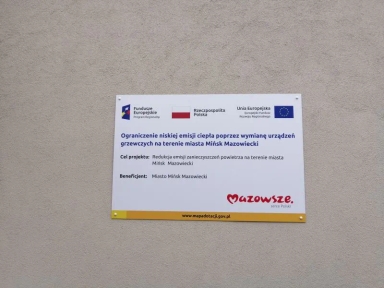 Tablica informacyjna o projekcie ograniczenia niskiej emisji ciepła poprzez wymianę urządzeń grzewczych na terenie miasta Mińsk Mazowiecki, z logotypami Funduszy Europejskich, Rzeczpospolitej Polski i Unii Europejskiej.