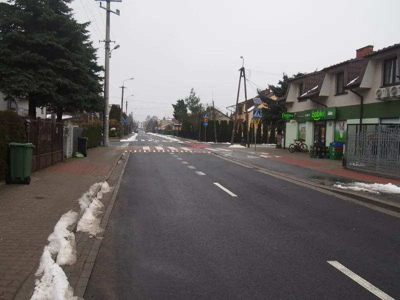 Pusta ulica z chodnikami po obu stronach, przejście dla pieszych i sklep Żabka po prawej stronie.