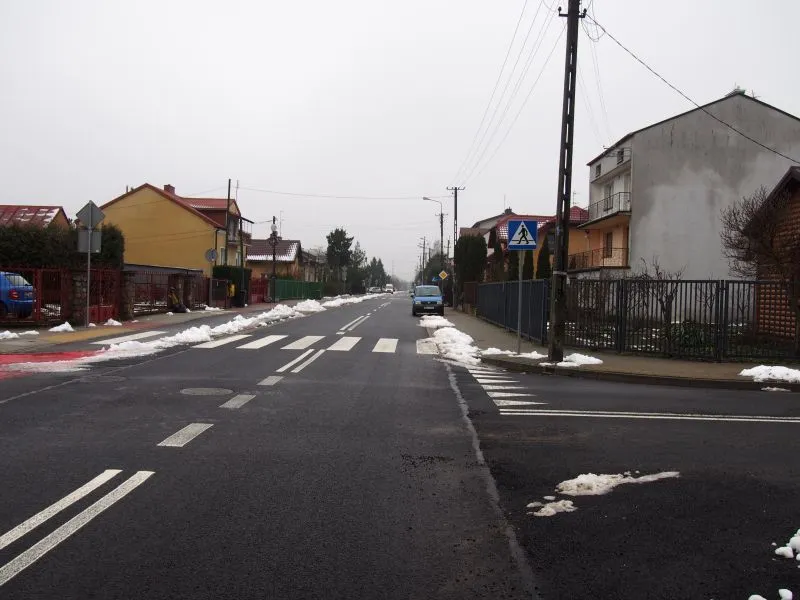 Ulica z namalowanym przejściem dla pieszych, zaparkowanym samochodem po prawej stronie oraz resztkami topniejącego śniegu na poboczach i chodnikach.