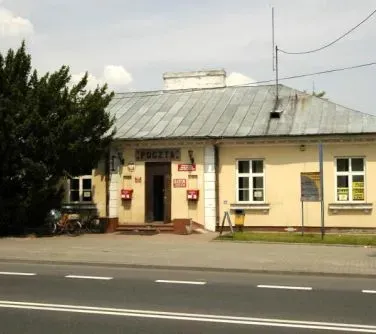 Budynek poczty z metalowym dachem, dwoma czerwonymi skrzynkami na listy przy wejściu i tabliczkami informacyjnymi.