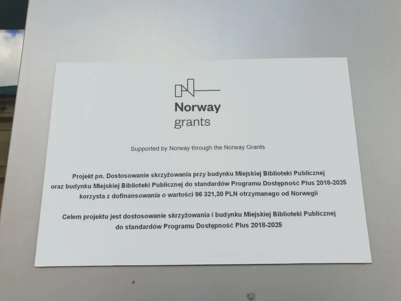 Tablica informacyjna o dofinansowaniu w wysokości 96 321,30 PLN od Norwegii na dostosowanie skrzyżowania i budynku Miejskiej Biblioteki Publicznej do standardów Programu Dostępność Plus 2018-2025.