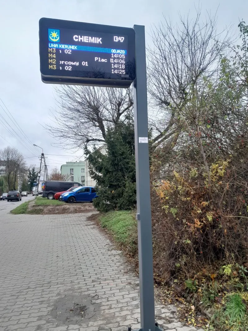 Elektroniczny tablica przystankowa z rozkładem jazdy autobusów i samochody zaparkowane obok chodnika.