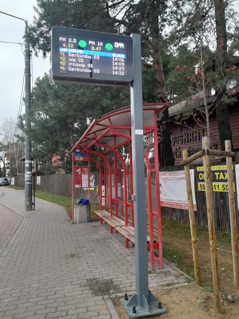 Przystanek autobusowy z czerwoną wiatą i elektroniczną tablicą wyświetlającą godziny odjazdów oraz poziom zanieczyszczenia powietrza.