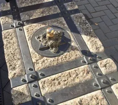 Metalowy emblemat osadzony na kamiennym bloku z metalowymi wzmocnieniami i nitami, znajdujący się na chodniku z kostki brukowej.