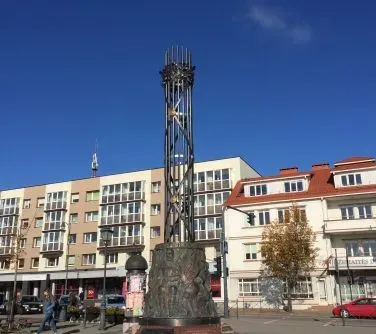 Stalowa rzeźba w formie wysokiej cylindrycznej konstrukcji z metalowymi prętami na cokole z płaskorzeźbami, ustawiona na miejskim placu obok budynków mieszkalnych.