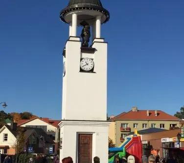 Biała wieża zegarowa z dzwonem na tle nieba i budynków mieszkalnych.