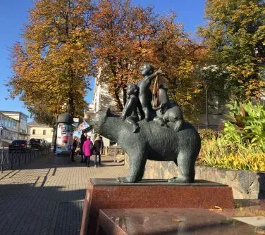Rzeźba niedźwiedzia z czterema dziećmi siedzącymi i stojącymi na jego grzbiecie, ustawiona na ceglanym postumencie przy chodniku.