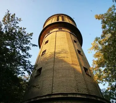 Wysoka, cylindryczna wieża wodna wykonana z cegły, otoczona drzewami.