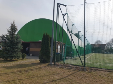 Po prawej boisko z zieloną murawą otoczone wysoką siatką i metalowymi słupami. Po lewej duża zielona kopuła za drewnianym budynkiem, przy wejściu stoją dwie osoby. Wokół rosną iglaste drzewa, niebo jest pochmurne.