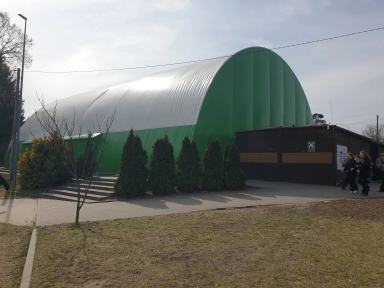 Duża zielono-biała hala namiotowa o zaokrąglonym dachu stoi na zewnątrz. Przed halą znajduje się rząd wysokich iglastych krzewów i małe bezlistne drzewko. Z lewej biegną schody prowadzące do wejścia. Po prawej stoi niska drewniana przybudówka, przy której idzie grupa osób. Niebo jest jasne, dzień.