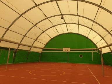 Hala sportowa pod białym, łukowym dachem z metalowymi żeberkami. Zielone ściany i czerwony kort z żółtymi liniami. Siatka do siatkówki rozciągnięta przez środek boiska, kosz zawieszony na tylnej ścianie.