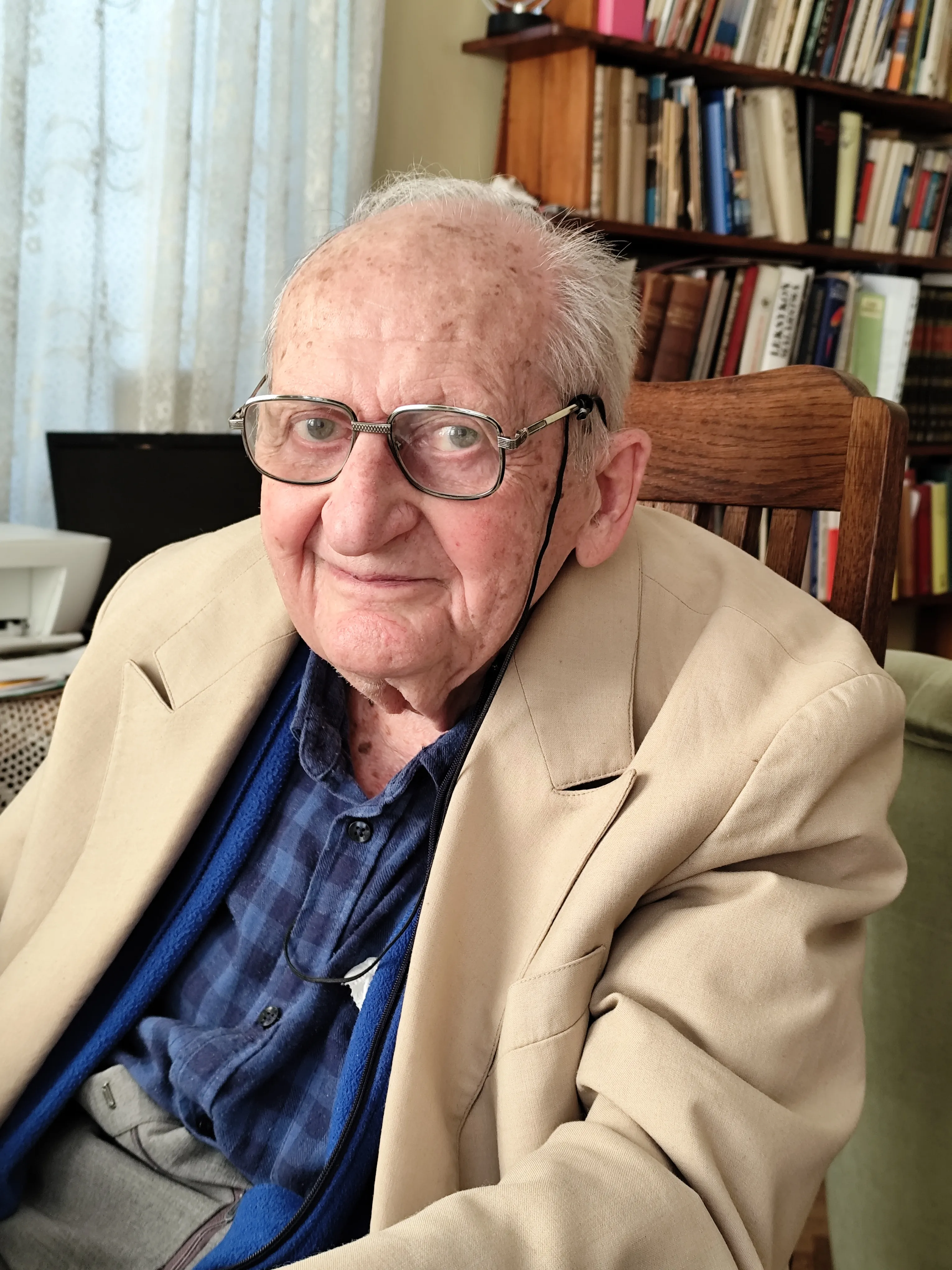 Kliknięcie powiększy obraz. Starszy mężczyzna w beżowej marynarce i okularach siedzi na drewnianym krześle w pokoju z regałem pełnym książek.