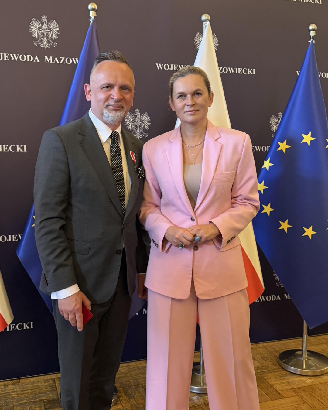 Kliknięcie powiększy obraz. Dwie stojące osoby na tle trzech flag, w tym flagi Unii Europejskiej i Polski. Mężczyzna w ciemnym garniturze i kropkowanej krawacie ma przypięty medal i trzyma mały czerwony przedmiot. Kobieta obok ma różowy garnitur i splecione dłonie. Tło jest ciemne z widocznymi godłami.