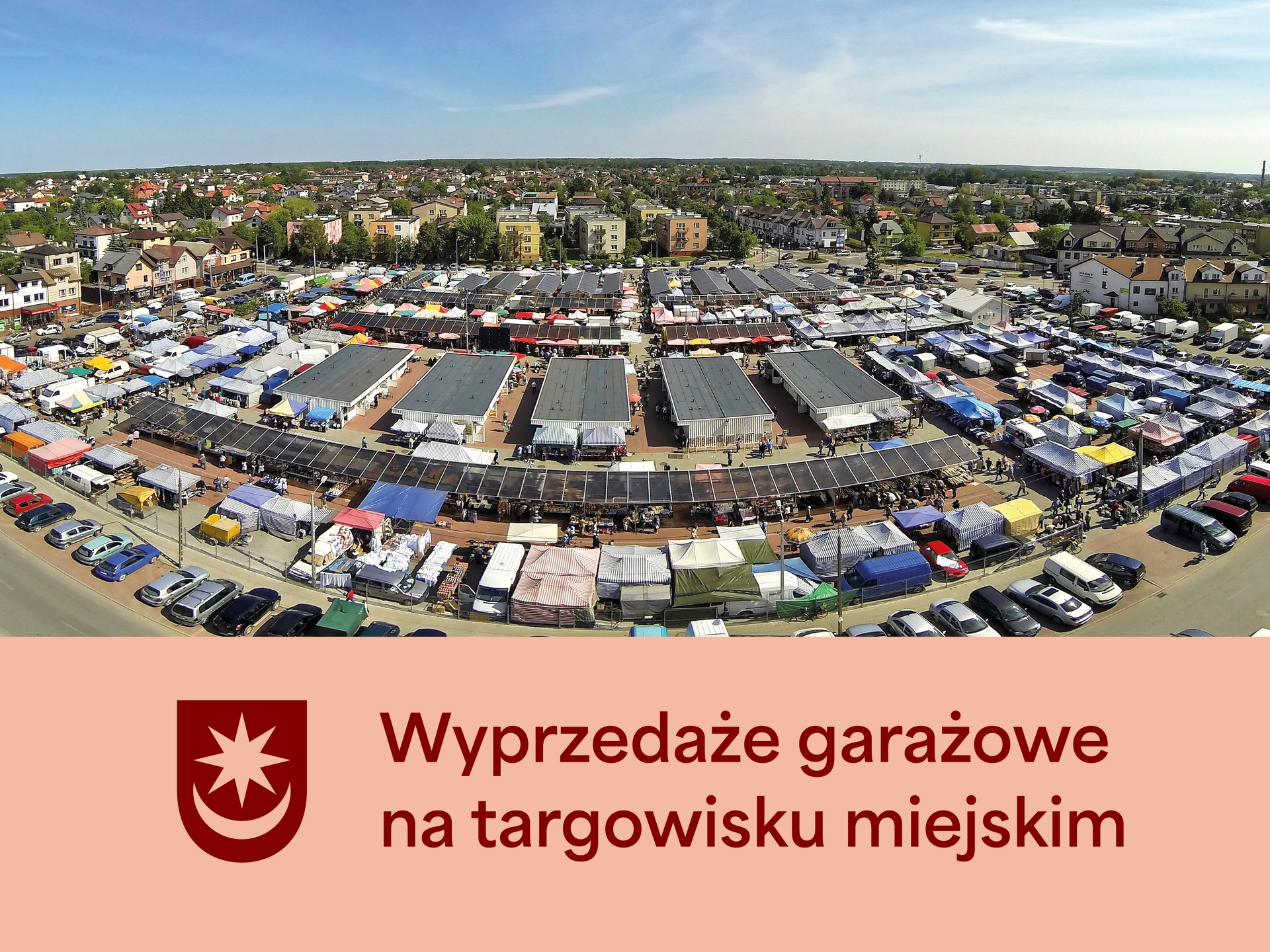 Kliknięcie powiększy obraz. Z lotu ptaka widać rozległe targowisko z rzędami zadaszonych stoisk i kolorowych namiotów, otoczone zaparkowanymi samochodami i niską zabudową mieszkalną pod jasnym, błękitnym niebem.