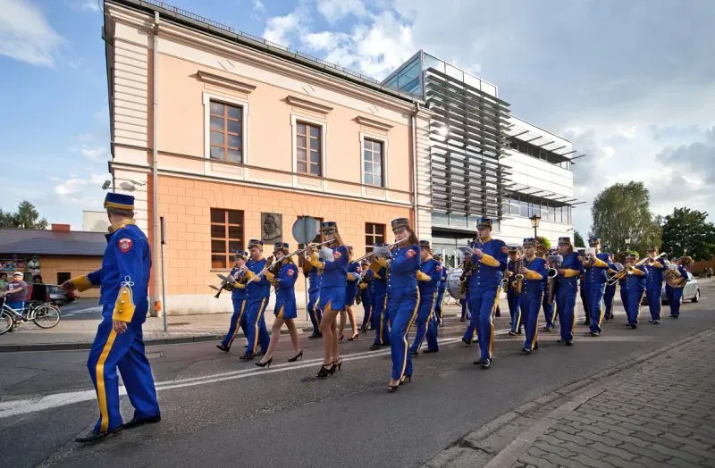 Grupa osób w niebieskich mundurach z żółtymi akcentami maszeruje ulicą grając na instrumentach dętych.