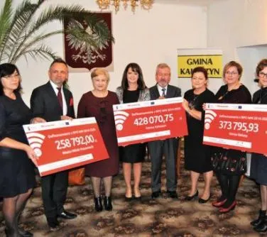 Kliknięcie powiększy obraz. Grupa osób stoi w pomieszczeniu, trzymając trzy duże czeki z widocznymi kwotami i nazwą „Gmina Kamień”.