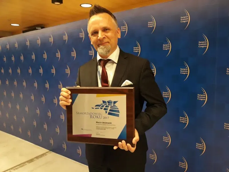 Mężczyzna w garniturze trzymający ramę z dyplomem "Samorządowiec Roku 2017" na tle niebieskiego banera z napisem "European Economic Congress".